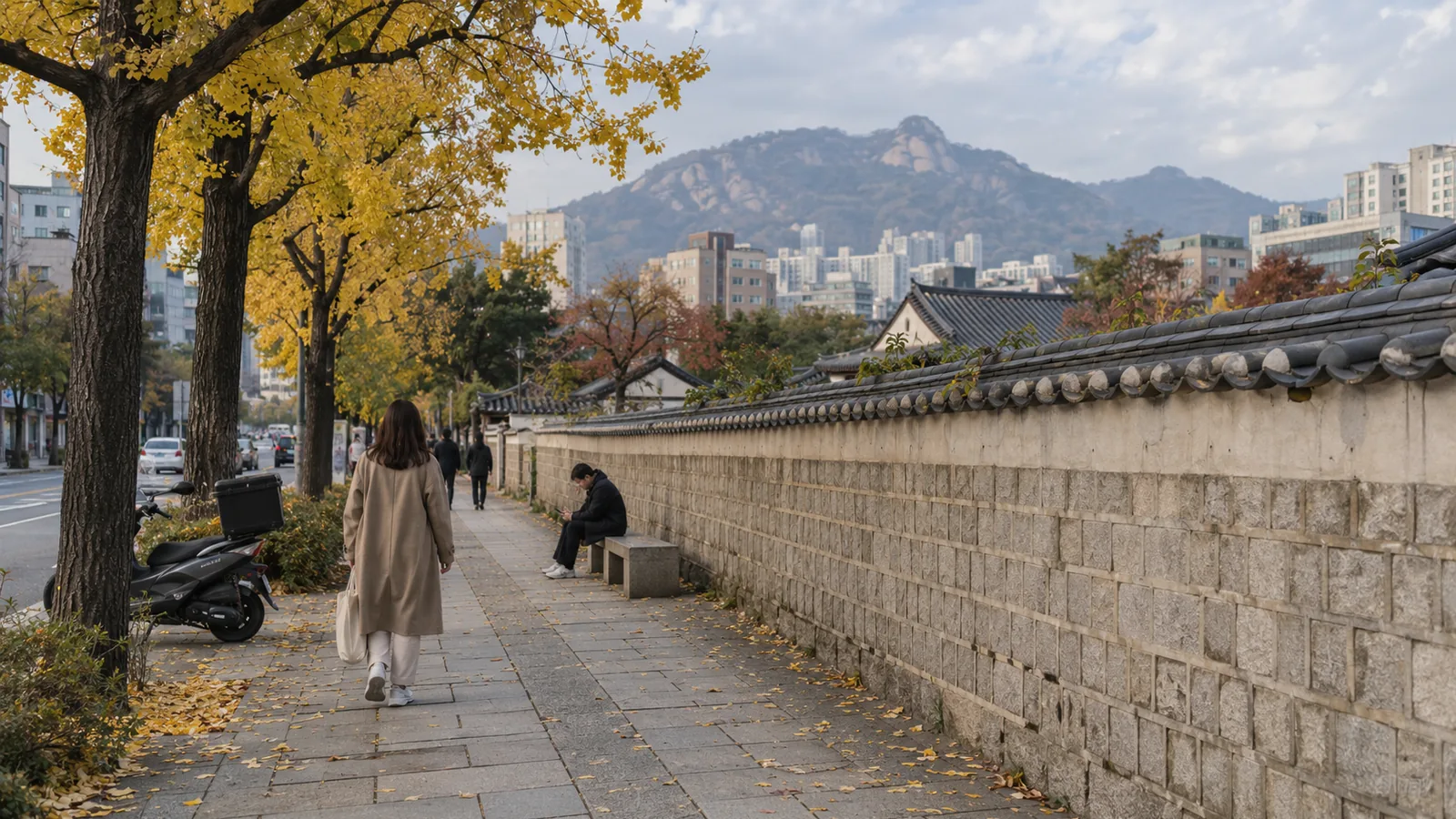 A woman walking past a tiled palace wall and yellow ginkgo trees on a quiet Seoul sidewalk in autumn