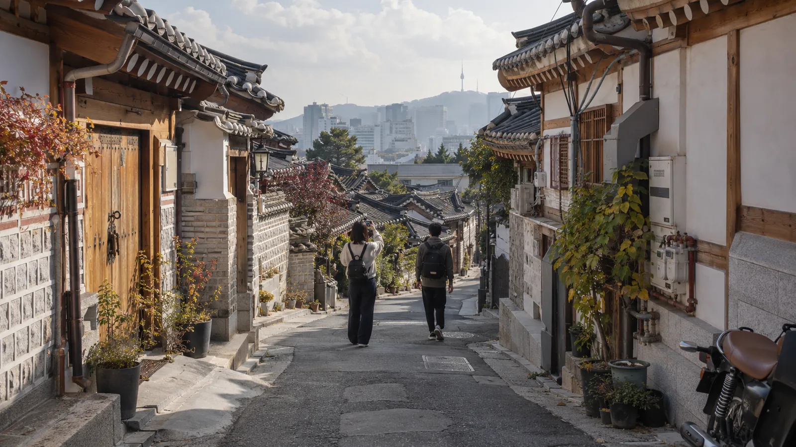 Two travelers walking up a hanok lane in Bukchon, Seoul, with N Seoul Tower on the hazy skyline